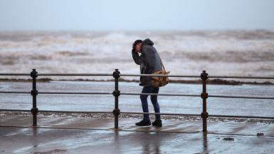 A man clutches his hat while walking along a windy seafront with rough waves in the background