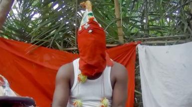A man wearing a red ceremonial mask covering his face sits in front of red and white clothes hung up in the bush to create a shrine for a juju practitioner in Sierra Leone. 