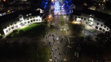 A drone shot of police forming a barricade while a group of people stand at the other end of a road. It is dark and they are lit by streetlights. 