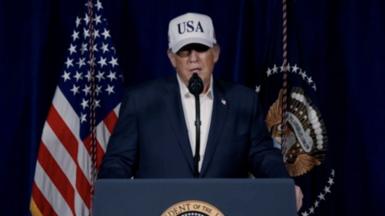 US President Donald Trump stands at a podium wearing a white cap with 'USA' written on it