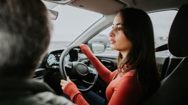 Stock image shows a young person turning a steering wheel as they reverse in a car while an instructor (out of frame) sits in the passenger seat.
