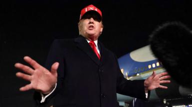 Donald Trump pictured from below with his hands outstretched, wearing a red USA baseball cap, with a plane visible in the background