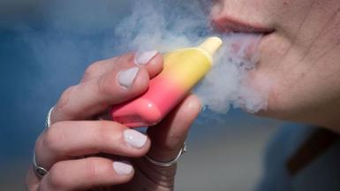 A woman holds a yellow and red coloured vape to her mouth with a cloud of vapour surrounding it.