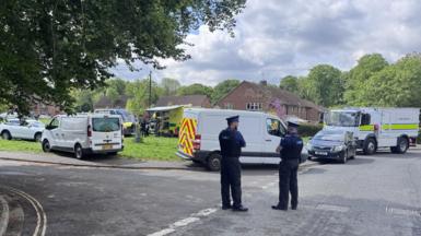 Two PCSOs are standing on a road, there are emergency vehicles in the middle of a patch of green grass