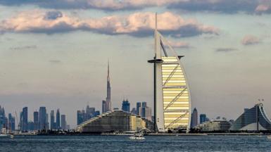 Buildings on the Dubai skyline, with tall towers seen in the background and the sail-shaped hotel the Burj Al Arab in the foreground, against a grey sky