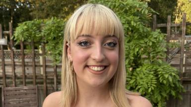 Imogen who has blonde mid length hair and a fringe looks at the camera smiling. Behind her is a brown garden fence and green leaves. 