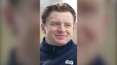 Adam Peaty looks to the side of the camera and smiles. He wears a navy jacket.