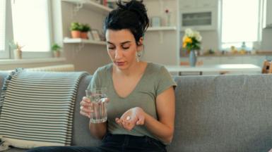 Young woman is sat with a glass of water in one hand and a white tablet in the open palm of the other. She is looking down so you can't see her eyes and her hair is tied up. She is sat on a grey sofa in what looks like an open plan living room with the kitchen behind her. 