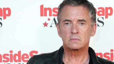 A man wearing a leather jacket and a black top standing in front of the press,
there is a backdrop behind him that reads inside soap awards 2024