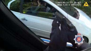 A close-up of someone's hands wearing black gloves, holding a black gun, pointing at a white car.