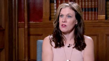 Natalie Fleet is sat down on a blue chair in front of a wooden bookcase. She is wearing a light pink top and has shoulder length hair.