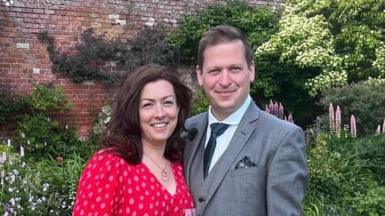 Ben and Sarah Howard standing in a garden. Sarah is wearing a red dress with an all-over spot pattern. She has brown hair. Ben is wearing a grey suit and dark tie