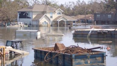 A flooded street in New Orleans with damaged houses, debris and an overturned vehicle