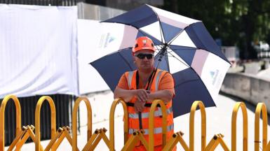 A worker in a high vis orange vest and helmet stands in the shade with a parasol above their head while standing next to a yellow barrier on a closed footpath in London on Tuesday - there is a sign next to him reading 'pedestrians please use other footpath'