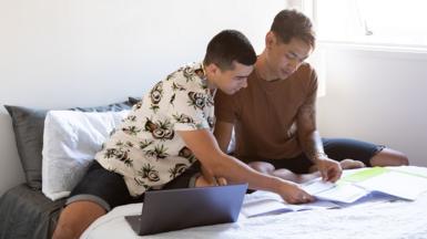 Stock photo shows a couple stiiting on a bed in their home looking at a laptop and bills.