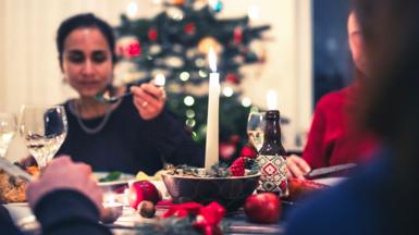 A person eats as part of a group during a festive meal, with a candle and festive food items on the table in the foreground and a Christmas tree in the background.