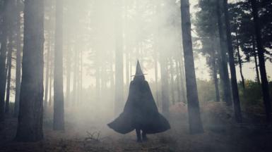 A woman with black cloak and a hat, walking through a misty forest