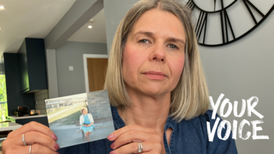 Michelle holds a small printed photograph with both hands. She is wearing a loose, long-sleeved top in a muted blue tone. The sleeves have gathered cuffs, and the fabric appears soft and casual. On her fingers, several rings are visible, including a band on one finger and another ring with a darker stone on a different finger. She is holding a small rectangular photograph at chest level. The photograph shows her mother sitting on a bench in sunlight, wearing a light blue outfit, with a building and grassy area in the background.