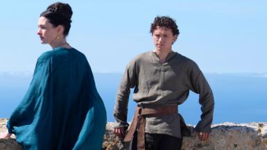 Tom Holland and Anne Hathaway in a film still from the film The Odyssey. Tom Holland is leaning against a wall with a scenic greek sea view behind him. He is wearing a dark olive green shirt and dark trousers. Anne hathaway is looking to the left and wearing a flowing flowing blue dress