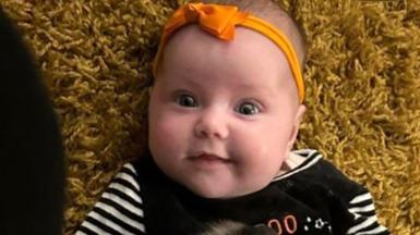 Dahlia-Rose Gartshore, a three-month-old baby girl looking at the camera, with a slight smile. She has an orange band on her head with a bow.