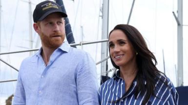 Harry and Meghan seen walking together in Sydney harbour in April. He is wearing a blue shirt and a cap, she is wearing a striped blue shirt.