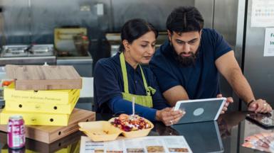 Stock photo shows two business owners looking at a tablet at the counter of a takeaway restaurant with food boxes on the counter and cooking appliances in the background.
