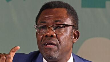 A close up shot of Veron Mosengo-Omba talking. He wears glasses and a blue suit.