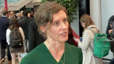 Jessica Toale is pictured giving an interview at the Labour party conference, with delegates in the background. She has tied-back brown hair and wears a green dress.