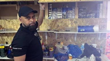 A person wearing a black shirt with 'ENFORCEMENT OFFICER' written on the sleeve is standing in front of wooden shelves. The person is lifting a wooden panel to reveal several stacks of boxes inside the shelf. Various items, including plastic containers, cleaning supplies, and other miscellaneous objects, are placed on the shelves below. A dog with black and white fur is visible at the bottom right corner of the image