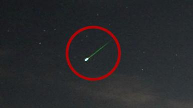 A meteor with a bright green tail streaks through the night sky, with several stars nearby. The meteor is circled with a red overlay.