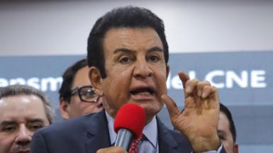 Presidential candidate Salvador Nasralla of Honduras' Liberal Party (PLH) gestures during a press conference. He wears a navy suit, blue short and red patterned tie. He is speaking into a bright red microphone