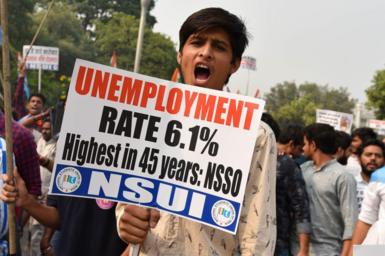 A young man wearing a white shirt holds a placard with slogans against unemployment and looks into the camera while shouting. Behind him are other protesters