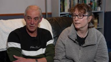 Ken and Kerstin Rudd. Ken is wearing a blue and white striped jumper and has short grey hair. Kerstin is wearing a fluffy grey top over a black t shirt and has glasses and short brown hair. Both are seated on a sofa.