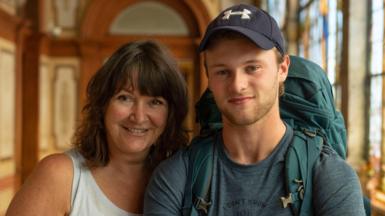 Jo and Sam pose together in an ornate room. He's wearing dark baseball cap and green top and rucksack and smiling, while Jo has shoulder-length brown hair and wears a white vest. She's also smiling.