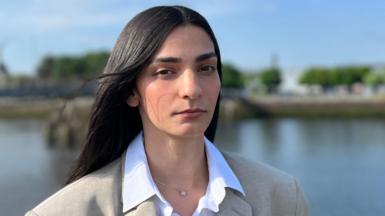 Head and shoulders shot of Ellie Wilson with a river in the background, which is out of focus. She wears a white shirt and an oversized grey blazer. She has long straight black hair.