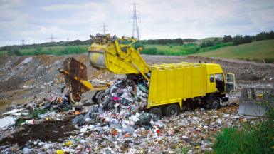 A landfill site having more waste dumped on it by a yellow truck. There are green hills in the background and electricity pylons can be seen dotting the horizon.