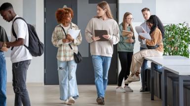 University students walking along a corridor towards the camera carrying books and work 