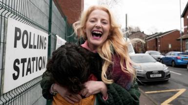 Hannah Spencer stands in front of a polling station sign attached to a metal fence. Spencer is smiling, holding an orange coloured bag and wearing a multi-coloured coat.