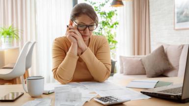 Woman in beige sweater with brown hair,wearing glasses looks at a pile of bills 