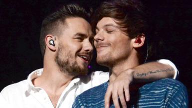 Liam Payne with his arm around and his face close to the face of a smiling Louis Tomlinson on stage