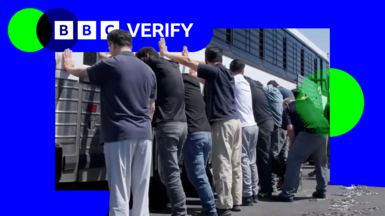A still frame from a video shows a group of men, with their backs to the camera and hands on the side of a white coach with black bars on its windows during an immigration raid at the Hyundai-LG vehicle assembly plant in Ellabell, Georgia. 
The men are in casual clothing, mostly jeans or slacks and T-shirts. 