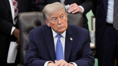 U.S. President Donald Trump sits at his desk in the Oval Office turning to his left. He is wearing a blue suit, white shirt and blue tie. He is not smiling. 