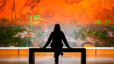 A silhouetted woman sitting on a gallery bench in front of Mohammed Sami's large painting