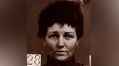 Black and white mugshot of a woman with dark hair piled on top of her head. She has a high collar around her neck and is looking slightly above the camera. There is a number - 28 - in the bottom left hand corner. 