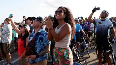 A woman smiles and claps as she watches the launch, with many people behind her looking to the skies, also watching.