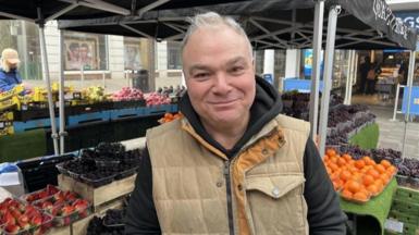 A man in a beige jacket smiling for the camera. Several varieties of fruit are stacked up in the background at a colourful fruit staff.