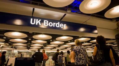 UK airport passport control. There is a blue overhead sign with UK border written on it in white. People are queuing to get their passports checked.