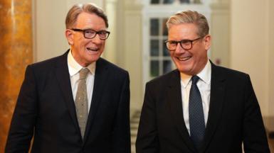 Lord Mandelson stands next to Keir Starmer. Both are wearing suits.