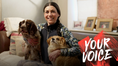 Helene Svinos sits on a sofa in between two dogs, with her arms around them. She is smiling and wearing a dark gilet over a patterned top, and leggings, with her dark hair tied back. There are cushions on the sofa and photo frames in the background.