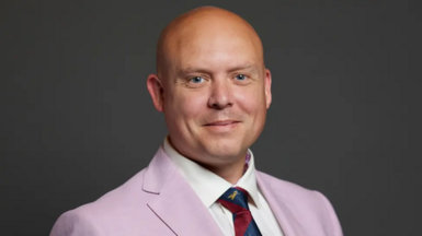 Head and shoulders shot of Cameron Thomas MP looking at the camera and smiling - wearing a pink suit, white shirt and red and blue stripped tie. 